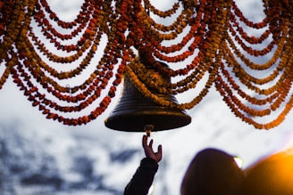 A hand reaches up to ring a large bell adorned with garlands of orange and yellow flowers. The background is slightly blurred, with suggestions of another person and what appears to be a snowy landscape.