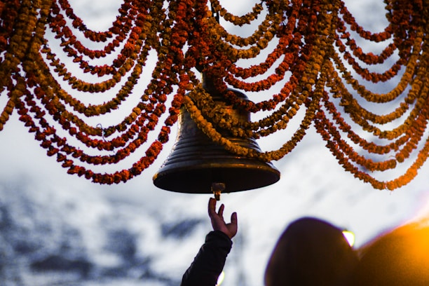 A hand reaches up to ring a large bell adorned with garlands of orange and yellow flowers. The background is slightly blurred, with suggestions of another person and what appears to be a snowy landscape.
