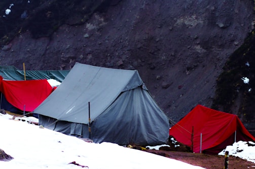 Several tents, primarily gray and red, are pitched on a snowy and rocky terrain with a rugged cliff face in the background.
