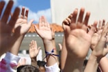 A group of raised hands reaching upwards, set against a sunny backdrop with a clear blue sky and a portion of a building visible. The hands show diversity in skin tones, and one has a blue bracelet. The image captures a sense of participation and unity.