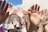 A group of raised hands reaching upwards, set against a sunny backdrop with a clear blue sky and a portion of a building visible. The hands show diversity in skin tones, and one has a blue bracelet. The image captures a sense of participation and unity.