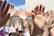 A group of raised hands reaching upwards, set against a sunny backdrop with a clear blue sky and a portion of a building visible. The hands show diversity in skin tones, and one has a blue bracelet. The image captures a sense of participation and unity.