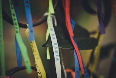 Colorful fabric ribbons hang from an object, each printed with text. The ribbons vary in color, including shades of blue, yellow, white, green, red, and purple. Some appear slightly blurred, while others are in focus against a soft, muted background.