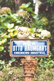A vintage street sign with ornate metalwork set against a lush green background. The sign reads 'Rua Otto Baumgart Engenheiro-Industrial'. The surroundings are vibrant with plants and flowers, giving a natural and historical feel.