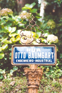 A vintage street sign with ornate metalwork set against a lush green background. The sign reads 'Rua Otto Baumgart Engenheiro-Industrial'. The surroundings are vibrant with plants and flowers, giving a natural and historical feel.