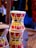 A wooden dholak or traditional drum with bright red, yellow, and green stripes is tightly bound with ropes, set against a blurred background that suggests a market or shop environment.