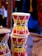 A wooden dholak or traditional drum with bright red, yellow, and green stripes is tightly bound with ropes, set against a blurred background that suggests a market or shop environment.