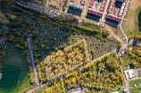 An aerial view of a landscape featuring a densely wooded area surrounded by a cemetery with visible tombstones arranged in neat rows. Beyond the cemetery, there is a construction site with several rectangular buildings with red roofs, and numerous vehicles and equipment. Adjacent to these, there are several green fields and patches of grass, with a few scattered trees. Roads with vehicles cut through the image, showing a mix of natural and urban environments.