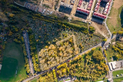 An aerial view of a landscape featuring a densely wooded area surrounded by a cemetery with visible tombstones arranged in neat rows. Beyond the cemetery, there is a construction site with several rectangular buildings with red roofs, and numerous vehicles and equipment. Adjacent to these, there are several green fields and patches of grass, with a few scattered trees. Roads with vehicles cut through the image, showing a mix of natural and urban environments.