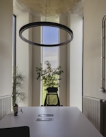 A modern office setting featuring a large circular light fixture hanging from the ceiling. A black office chair is placed at the end of a white conference table, with two slots embedded in the table's surface. The room is brightened by vertical windows, revealing a view of lush greenery outside. Potted plants are positioned near the windows, adding a touch of nature to the space. Radiators are installed along the walls, and a few office supplies are placed on the table.