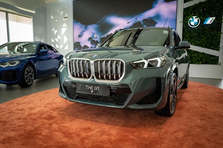 A sleek, modern BMW SUV is prominently displayed indoors on a circular, plush orange carpet. The vehicle has a shiny, metallic finish and distinctive grille design. In the background, a blue BMW sedan is also visible to the left. The setting includes a large screen with an abstract image and green plant decorations on the walls, adding a contemporary and elegant feel to the environment.