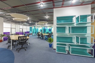 A modern office space with open seating arrangements, featuring several desks with rolling chairs. The ceiling has a unique geometric design with hanging light fixtures. Bright blue shelves are mounted on a wall, and potted plants are scattered around, adding a touch of nature. Large windows on one side allow natural light to illuminate the area.