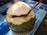 A fresh green coconut with the top partially cut open sits on a floral-patterned plate. A red straw is inserted into the coconut, which rests on a table with a blue geometric pattern.
