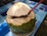 A fresh green coconut with the top partially cut open sits on a floral-patterned plate. A red straw is inserted into the coconut, which rests on a table with a blue geometric pattern.