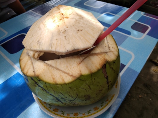 A fresh green coconut with the top partially cut open sits on a floral-patterned plate. A red straw is inserted into the coconut, which rests on a table with a blue geometric pattern.