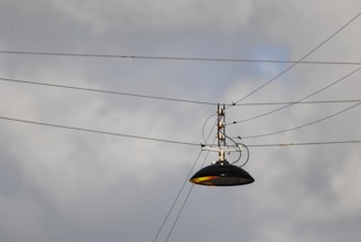 A streetlight suspended by several cables against a cloudy sky. The lamp is metallic and dark in color, with multiple wires attached to its metal frame, creating an industrial feel.