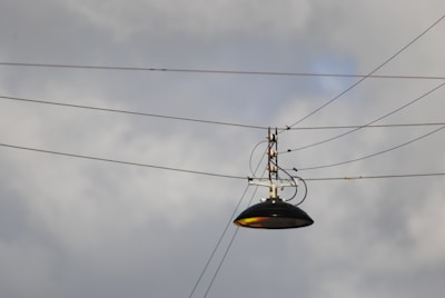 A streetlight suspended by several cables against a cloudy sky. The lamp is metallic and dark in color, with multiple wires attached to its metal frame, creating an industrial feel.