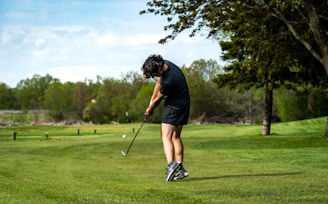 A golfer swinging a club on a lush green golf course under a clear blue sky.