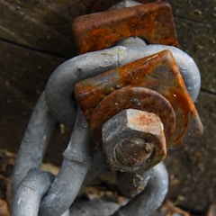 A close-up view of a heavily rusted bolt and nut on a chain link. The metallic surfaces show signs of significant corrosion with an orange-brown hue. The background appears to be a wooden surface.