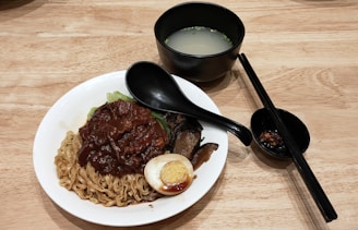 A wooden table holds a white bowl filled with noodles, stewed meat, leafy greens, a halved boiled egg, and mushrooms. A black spoon rests on the bowl. A separate black bowl contains a clear broth with green garnishes. Next to it, a small black dish holds chili paste and a pair of black chopsticks.
