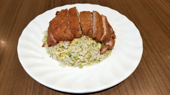 Crispy slices of fried chicken laid over a mound of fried rice with chopped vegetables and egg on a white plate. The dish is presented on a wooden surface.