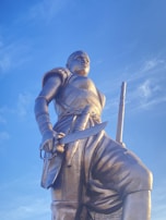 A large bronze statue of a warrior holding a sword, dressed in detailed armor with bands and patterned motifs. The figure stands against a clear blue sky, exuding strength and determination. The craftsmanship highlights intricate details in the armor and weaponry.
