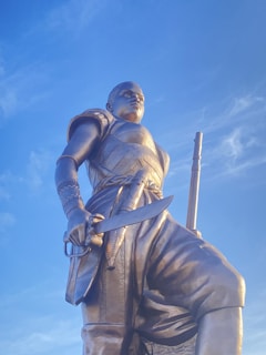 A large bronze statue of a warrior holding a sword, dressed in detailed armor with bands and patterned motifs. The figure stands against a clear blue sky, exuding strength and determination. The craftsmanship highlights intricate details in the armor and weaponry.