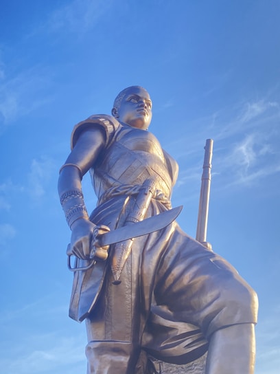 A large bronze statue of a warrior holding a sword, dressed in detailed armor with bands and patterned motifs. The figure stands against a clear blue sky, exuding strength and determination. The craftsmanship highlights intricate details in the armor and weaponry.