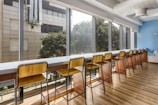 A long row of wooden bar stools is lined up against a bar-height counter next to large windows, allowing natural light to flood into a modern interior space. The wooden floor complements the furniture, and the blue accent wall adds a touch of color. Outside the windows, trees and a building facade are visible.