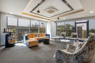 A modern lounge area with large windows offering a view of the outdoors. The space features a mix of seating options, including a blue and tan sectional sofa, patterned armchairs, and gray cushioned chairs. A small bar area with various bottles is placed near the window. The ceiling has contemporary lighting and an air conditioning unit. The colors and decor create a stylish and comfortable environment.