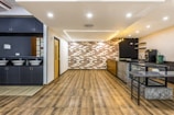A modern and spacious interior with a sleek design, featuring wooden flooring and a combination of dark and light color tones. The room includes a small kitchenette area with overhead lights, a counter with a green bag and appliances like a coffee machine. On the left, there are sinks with mirrors and grey cabinetry.