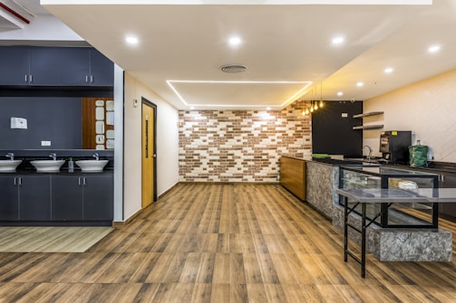A modern and spacious interior with a sleek design, featuring wooden flooring and a combination of dark and light color tones. The room includes a small kitchenette area with overhead lights, a counter with a green bag and appliances like a coffee machine. On the left, there are sinks with mirrors and grey cabinetry.