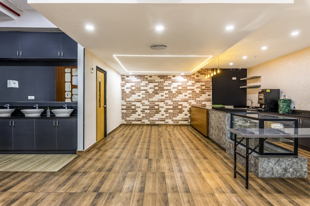 A modern and spacious interior with a sleek design, featuring wooden flooring and a combination of dark and light color tones. The room includes a small kitchenette area with overhead lights, a counter with a green bag and appliances like a coffee machine. On the left, there are sinks with mirrors and grey cabinetry.
