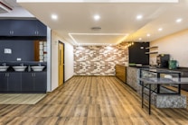 A modern and spacious interior with a sleek design, featuring wooden flooring and a combination of dark and light color tones. The room includes a small kitchenette area with overhead lights, a counter with a green bag and appliances like a coffee machine. On the left, there are sinks with mirrors and grey cabinetry.