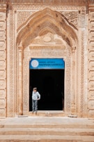An ornate archway with intricate carvings frames the entrance to a building, with a sign above the door that reads 'Gazipasa Ilkokulu' indicating it is a primary school in Mardin, Artuklu. A child stands in the doorway, looking to the side, wearing a light-colored shirt and pants.