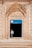 An ornate archway with intricate carvings frames the entrance to a building, with a sign above the door that reads 'Gazipasa Ilkokulu' indicating it is a primary school in Mardin, Artuklu. A child stands in the doorway, looking to the side, wearing a light-colored shirt and pants.