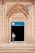 An ornate archway with intricate carvings frames the entrance to a building, with a sign above the door that reads 'Gazipasa Ilkokulu' indicating it is a primary school in Mardin, Artuklu. A child stands in the doorway, looking to the side, wearing a light-colored shirt and pants.