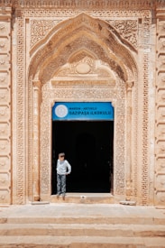 An ornate archway with intricate carvings frames the entrance to a building, with a sign above the door that reads 'Gazipasa Ilkokulu' indicating it is a primary school in Mardin, Artuklu. A child stands in the doorway, looking to the side, wearing a light-colored shirt and pants.