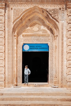 An ornate archway with intricate carvings frames the entrance to a building, with a sign above the door that reads 'Gazipasa Ilkokulu' indicating it is a primary school in Mardin, Artuklu. A child stands in the doorway, looking to the side, wearing a light-colored shirt and pants.