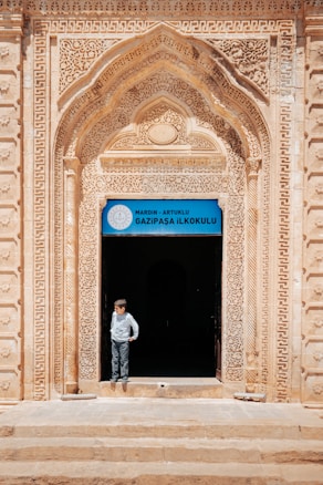 An ornate archway with intricate carvings frames the entrance to a building, with a sign above the door that reads 'Gazipasa Ilkokulu' indicating it is a primary school in Mardin, Artuklu. A child stands in the doorway, looking to the side, wearing a light-colored shirt and pants.