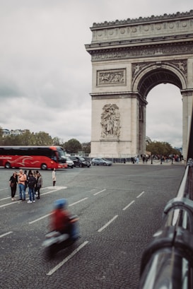 A busy urban scene featuring a famous historical monument with intricate carvings. Several people are gathered on the sidewalk, some appearing to be tourists. A red tour bus and several cars are visible on the road, and a person on a scooter is seen in motion, creating a sense of movement.