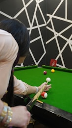 A person is poised to take a shot on a green snooker or pool table. The player, seen from behind, holds a cue stick aimed at several colored balls on the table. The background features a geometric black and white pattern, adding an artistic touch to the scene.