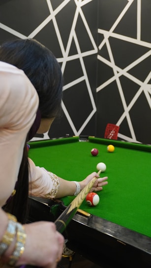 A person is poised to take a shot on a green snooker or pool table. The player, seen from behind, holds a cue stick aimed at several colored balls on the table. The background features a geometric black and white pattern, adding an artistic touch to the scene.