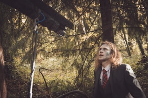 A person with long hair and a pale face is wearing a formal suit and a red plaid tie, standing in a forest setting. The individual is looking upwards, and there's a large microphone boom visible, suggesting a film or video production environment.