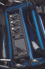 A portable audio mixer or recording device is nestled inside a protective case with blue foam padding. The device features multiple control knobs and buttons, a digital display screen, and various input ports. The surroundings include cables and metal hooks.