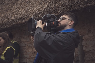 A person with a beard and glasses operates a professional video camera outdoors. The individual is wearing a dark jacket with a blue scarf and seems focused on filming. The background consists of a textured wall and a thatched roof, giving a rustic atmosphere. Another person in a yellow vest is partially visible.