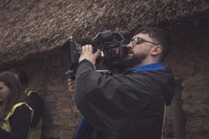 A person with a beard and glasses operates a professional video camera outdoors. The individual is wearing a dark jacket with a blue scarf and seems focused on filming. The background consists of a textured wall and a thatched roof, giving a rustic atmosphere. Another person in a yellow vest is partially visible.