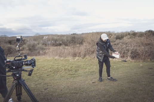A person wearing a white cap and black winter clothing is standing on a grassy field holding a clapperboard. A professional camera setup with a tripod and monitor is positioned nearby, aimed towards the person. The background features a stretch of barren trees and cloudy skies, suggesting an outdoor film set scene.