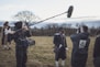 A film crew is on set in an outdoor grassy field. The central figure is dressed in period costume, wearing a dark coat, hat, and white stockings, while speaking to the camera. A sound technician holds a boom microphone, and a cameraman captures the scene. Another person in period costume stands in the background, with bare trees and a cloudy sky surrounding the set.