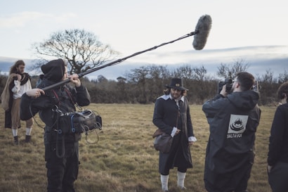 A film crew is on set in an outdoor grassy field. The central figure is dressed in period costume, wearing a dark coat, hat, and white stockings, while speaking to the camera. A sound technician holds a boom microphone, and a cameraman captures the scene. Another person in period costume stands in the background, with bare trees and a cloudy sky surrounding the set.